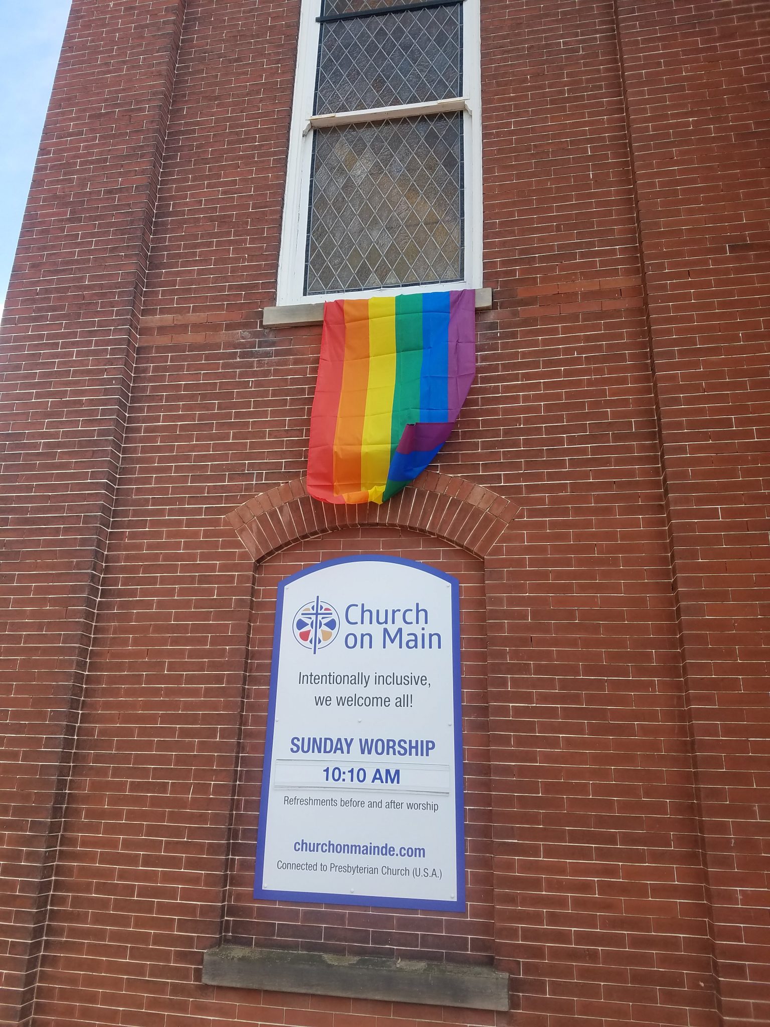 A picture of the front of the church. A LGBTQ pride flag is hanging from a window. A Sign on the front of the building states "Church on Main, Intentionally Includsive, We welcome all. Sunday Worship 10:10AM. Refreshments before and after worship (Outdated on the sign but included here for accuracy.) Churchonmainde.com. Connected to the Presbyterian Church. (U.S.A.)